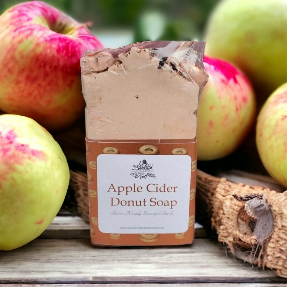 A bar of Apple Cider Donut Soap with a beige to brown gradient color, packaged with a label in front, set against a backdrop with apples and apple cider.