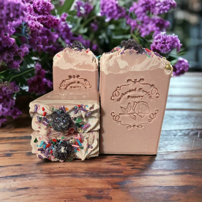 Three bars of soap with intricate tops, placed on a wooden surface with purple flowers in the background.